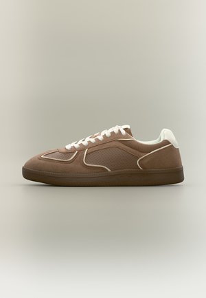 Bruine suède sneaker met witte veters en afwerking, geperforeerd zijpaneel en donkerbruine zool op een neutrale achtergrond.