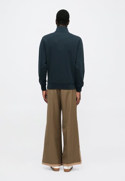Felpa blu navy con collo alto, abbinata a pantaloni larghi color kaki con fessure laterali e finitura testurizzata, mostrati da dietro.