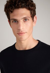 Schwarzer Rundhalsausschnitt-Pullover mit strukturiertem Strick. Das Modell hat kurze, lockige Haare und einen neutralen Hintergrund, der die Details der Kleidung hervorhebt.