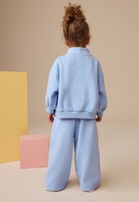 Lyseblå fleecetrøje med krave og ribbede manchetter, parret med matchende vide bukser, sat mod en neutral baggrund.