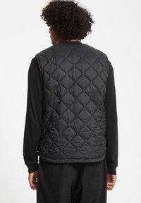 Sort quiltet vest med diamantmønster, lavet af letvægtsmateriale. Har en rund hals og en glat tekstur, båret over en sort langærmet skjorte.