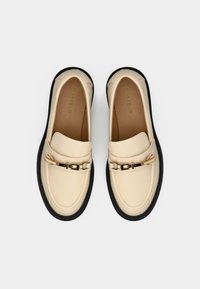 Paire de mocassins en cuir beige avec semelles noires et détails de boucles dorées, vus de dessus sur fond blanc.