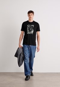 Zwarte T-shirt met korte mouwen en een grafische print op de voorkant, gecombineerd met blauwe spijkerbroek en zwarte schoenen, terwijl er een grijze jas wordt vastgehouden.