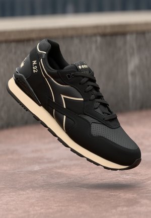 Scarpa sportiva nera realizzata in materiali sintetici. Presenta una finitura testurizzata, una suola color crema e sottili accenti beige con sezioni perforate.