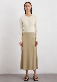 Cardigan beige clair avec des boutons, associé à une jupe longue et lisse couleur kaki. Les chaussures consistent en des sandales à glissière marron avec une boucle.