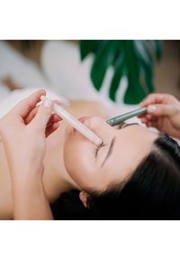 Dwa gładkie aplikatory, jeden biały i jeden zielony, są używane blisko nosa osoby podczas zabiegu kosmetycznego. W tle widać rozmyte liście.