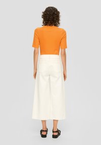 Pomarańczowy dopasowany t-shirt z krótkimi rękawami w połączeniu z białą, szeroką spódnicą dżinsową. Widoczne czarne sandały na platformie. Minimalistyczny design.