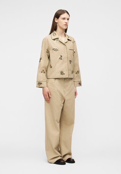 Γυναίκα που φοράει μπεζ ριγέ πουκάμισο με κεντήματα από λουλούδια και μπεζ παντελόνι φαρδιά με διακριτικά πουά σχέδια, στέκεται μπροστά σε απλό φόντο.