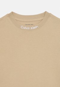 Beige cotton t-shirt with a crew neckline. Features "Calvin Klein Jeans" logo in white embroidery along the neckline. Soft texture.