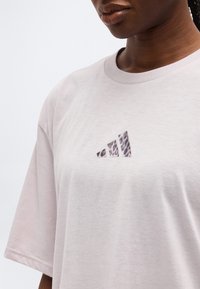 Κοντινό πλάνο ενός ατόμου που φοράει ένα ανοιχτό ροζ μπλουζάκι με το μοτίβο του λογότυπου Adidas στο κέντρο του στήθους.