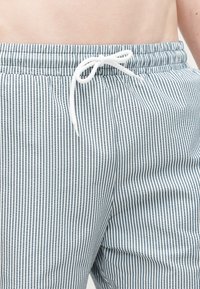 Gros plan sur la taille d'une personne portant un short à cordon à rayures verticales bleues et blanches, noué avec un nœud blanc.