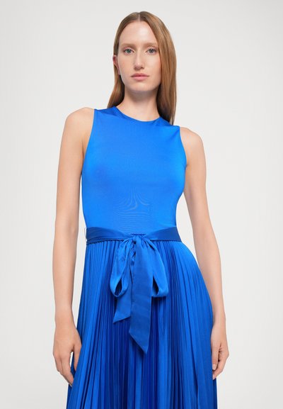 Frau mit glattem roten Haar, die ein ärmelloses, leuchtend blaues Kleid mit einem plissierten Rock und einer satinierten Schleife in der Taille trägt.