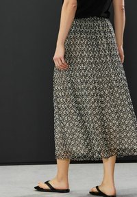 Maxi-hame, joka esittelee musta-beige-geometrisia kuvioita, pleatattua muotoilua, joustavaa vyötäröä ja kevyttä materiaalia yhdistettynä mustiin sandaaleihin.