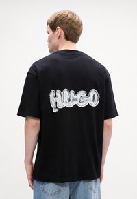 Černé bavlněné tričko s velkým, lesklým bílým logem "HUGO" na zadní straně. Krátké rukávy a uvolněný střih s kulatým výstřihem.