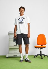 Camiseta blanca de manga corta con estampado gráfico negro, combinada con pantalones cortos oscuros. Zapatillas grises con toques blancos y suelo de césped verde.