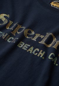 Marine t-shirt med et flerfarvet rhinestone grafik, der stavar "SuperDry" og "Venice Beach, CA." Blødt stof med rund hals.
