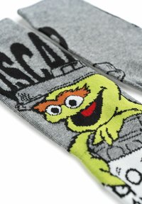 Calzini grigi con un personaggio verde dai capelli arancioni e dettagli neri, su uno sfondo grigio con un design di un cestino dell'immondizia.