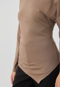 Langermet topp i taupe, laget av mykt, elastisk stoff. Har en buet søm og slank passform, kombinert med svarte bukser.
