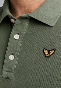 Zelené polokošile vyrobené z texturované tkaniny s límcem, s bílými knoflíky a černým křídletem se zlatým akcentem na levé straně.