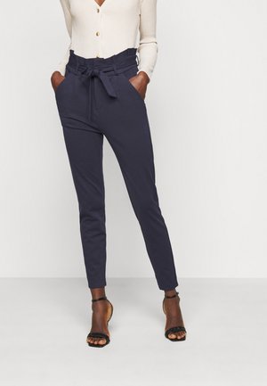 Frau trägt marineblaue Paperbag-Hosen, cremefarbenes geripptes Oberteil mit Knöpfen und schwarze Knöchelriemen-Absätze, steht mit Händen in den Taschen.