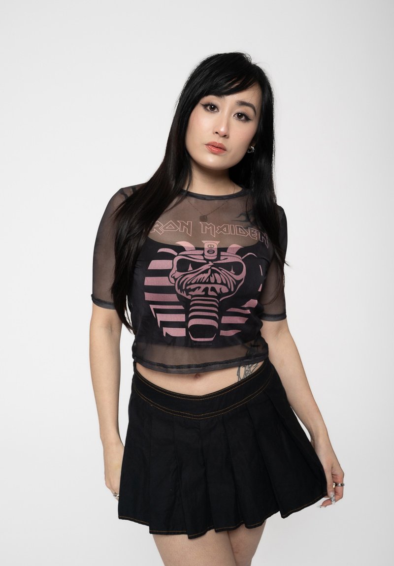 Black mesh crop top featuring a pink Iron Maiden graphic design, paired with a black pleated skirt. Smooth texture and fitted style.