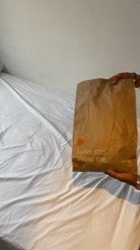 Καφέ χαρτοσακούλα κρατημένη πάνω από ένα λευκό σεντόνι, με εκτυπωμένο κείμενο "Αγάπα με. Φόρεσέ με." και ένα πορτοκαλί γραφικό καρδιάς. Το κείμενο είναι λευκό.