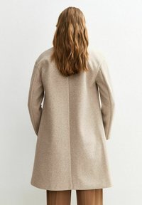 Manteau en laine beige avec une silhouette droite, des manches généreuses et une couture centrale à l'arrière. La texture semble douce et chaude.