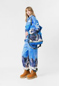 Abrigo y pantalones de estampado azul con imágenes de montañas, combinados con un bolso azul texturizado de gran tamaño. Botas marrones con suelas gruesas.