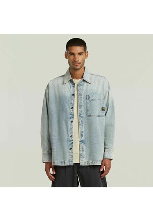 Uomo che indossa una giacca in denim azzurro chiaro sopra una camicia beige e pantaloni neri, in piedi davanti a uno sfondo chiaro uniforme.