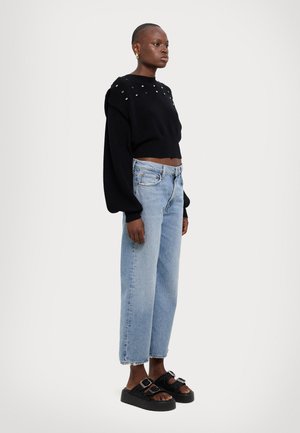 Black cropped sweater with studded details on shoulders, paired with light blue high-waisted wide-leg jeans and chunky black sandals.