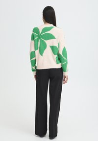 Cremefarbener Pullover mit großen grünen Blattmustern, gefertigt aus weicher Wolle, kombiniert mit schwarzen, weit geschnittenen Hosen. Minimalistisches Design, keine sichtbaren Metallteile.