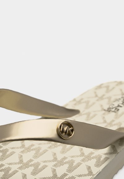 Κοντινό πλάνο μπεζ σαγιονάρας με μεταλλικό λουράκι και στρογγυλό λογότυπο MK, με σχεδιασμένο πέλμα με επαναλαμβανόμενα γράμματα MK σε ανοιχτό καφέ.