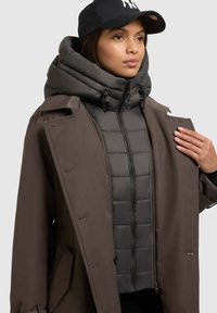 Cappotto esterno marrone con dettagli in bottoni, sovrapposto a un giubbotto imbottito grigio. Presenta un grande cappuccio imbottito e un berretto nero.