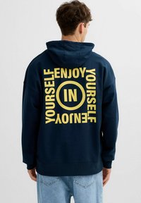 Hombre con sudadera con capucha azul marino con texto amarillo "ENJOY YOURSELF" y "IN" dentro de un círculo en la parte trasera, combinado con vaqueros azul claro.