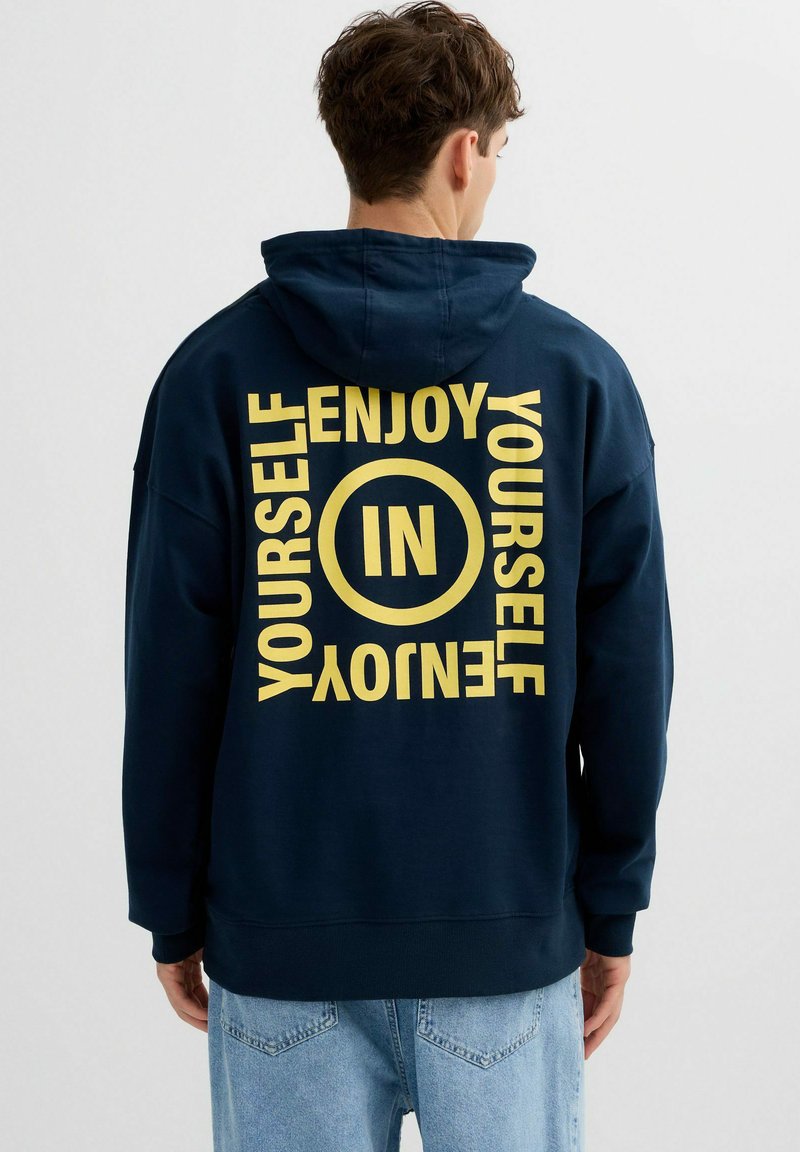 Hombre con sudadera con capucha azul marino con texto amarillo "ENJOY YOURSELF" y "IN" dentro de un círculo en la parte trasera, combinado con vaqueros azul claro.