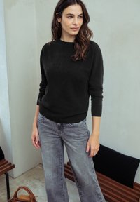 Žena s dlouhými hnědými vlasy, oblečená v černém svetr a šedých džínech, stojí blízko dřevěné lavičky a hnědého kabelky na dlažbě.