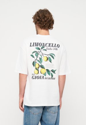 Homme aux cheveux bouclés portant un T-shirt blanc avec une branche de citronnier et le texte "Limoncello Dolce Vita La Gioia di Vivere" dans le dos.