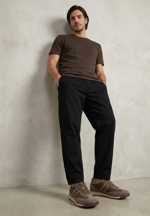 Mand står op ad en struktureret lys grå væg iført en brun t-shirt, sorte bukser og beige og brune sneakers med striber.