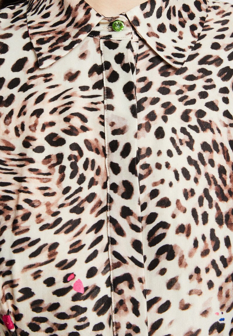 Blouse à imprimé léopard en tissu doux et léger, avec un col classique et un bouton vert au niveau du cou. Des accents de rose rehaussent le design.
