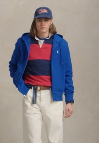 Moletom azul com fecho éclair sobre uma camisa polo às riscas vermelha e azul-marinho, calças de cor creme e um boné azul. Apresenta um cinto às riscas e um logótipo branco.