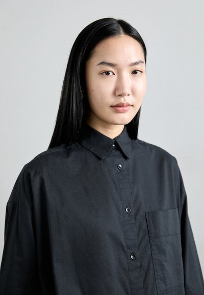 Black button-up shirt in a lightweight, textured fabric. Features a collar, front pocket, and black buttons. Simple, oversized design.