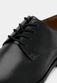 Zapato negro de cuero con cordones, acabado liso, puntera redondeada, seis ojales y costuras en contraste en el lateral. Presenta cordones delgados negros.