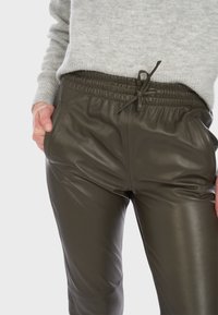 Olivově zelené kožené joggery s elastickým pasem, zavazovací šňůrkou a bočními kapsami. Hladká textura a přiléhavý střih.