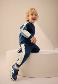 Granatowa bluza i dresy z kremowymi i jasnoniebieskimi akcentami. Dziecięce buty sportowe są granatowe z białą podeszwą i czarnymi sznurówkami.