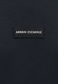 Μαύρο υφασμάτινο σχέδιο με μια μικρή ορθογώνια ραμμένη ετικέτα που εμφανίζει την επιγραφή "ARMANI EXCHANGE" με λευκά κεφαλαία γράμματα.
