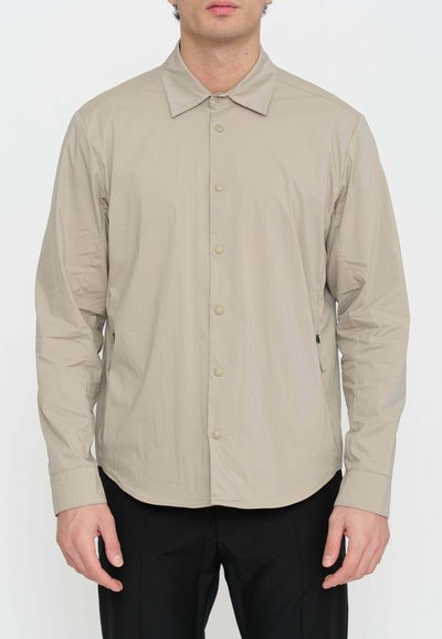 Camicia beige con colletto a punta, maniche lunghe e texture liscia. Presenta bottoni frontali e tasche laterali con zip.