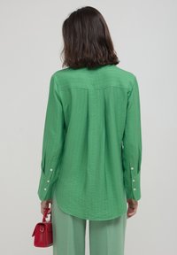 Woman seen from behind wearing a long-sleeve green striped shirt and pants, holding a small red handbag in her right hand.