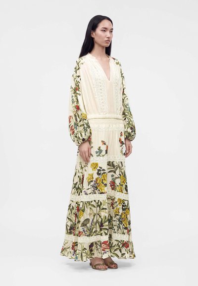 Femme debout portant une longue robe crème avec des motifs floraux et des détails en dentelle, associée à des sandales à bout ouvert sur un fond uni.