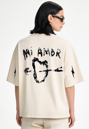Frau trägt ein beige oversized T-Shirt mit dem Text "Mi Amor", einem von einem Pfeil durchbohrten Herz auf dem Rücken und schwarzen Blitz-Designs auf den Ärmeln.