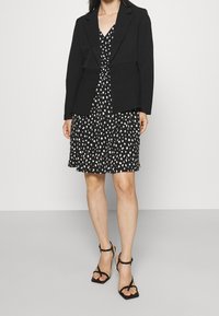 Černý blazer přes šaty s černobílým vzorem a výstřihem do V. Outfit doplňují černé sandály na podpatku. Hladká tkanina a strukturovaný střih.